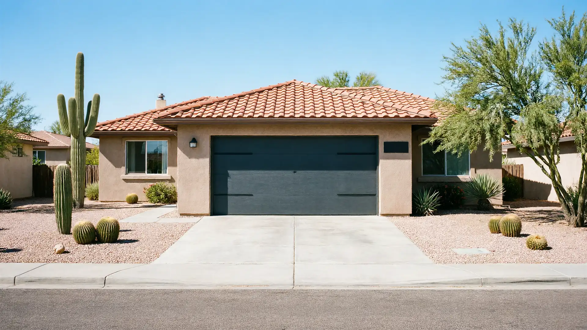 The house has a dark gray garage door, a tiled roof, and desert landscaping.